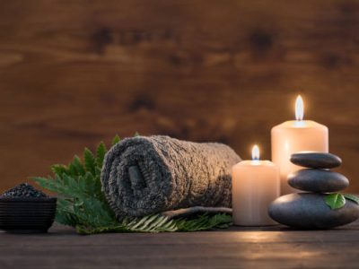 Brown towel on green fern with candles and black hot stone on wooden background. Hot stone massage setting lit by candles. Hot stone therapy for one person with candle light. Beauty spa treatment and relax concept.